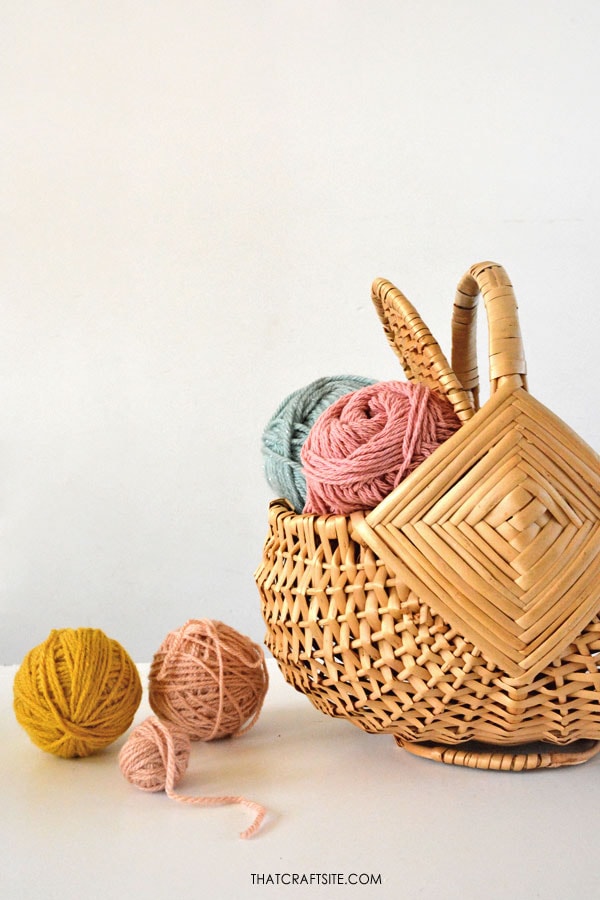 Vintage wicker basket with pink and light blue yarn in it. Next are three smaller yarn balls in mustard yellow and pale pink. Text on photo: thatcraftsite.com.