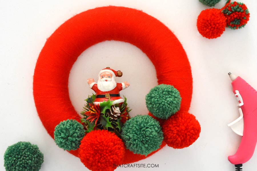 Step 6 how to make a yarn wrapped Christmas wreath. Position and attach your pom poms and other decorative items. thatcraftsite.com