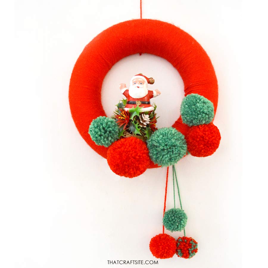 Step 7 how to make a yarn Christmas pom pom wreath. Add pom poms to the bottom and a string of yarn to hang your finished wreath. thatcraftsite.com