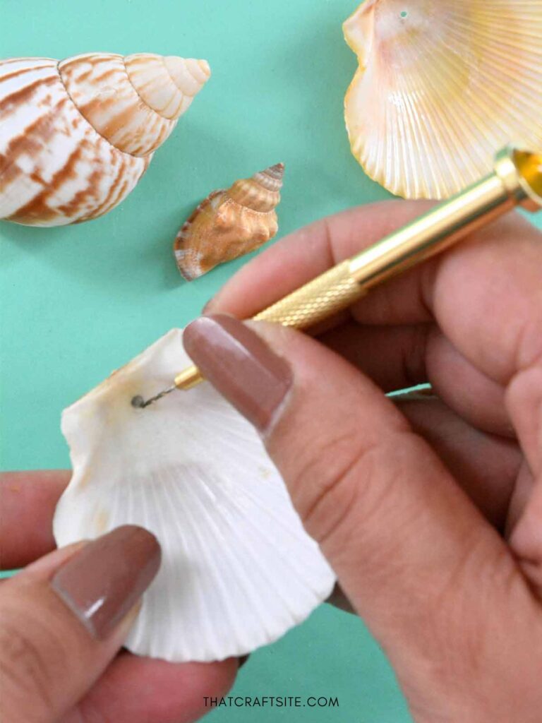 Female hands drilling hole in seashell with a gold colored hand drill. More shells in the background.
