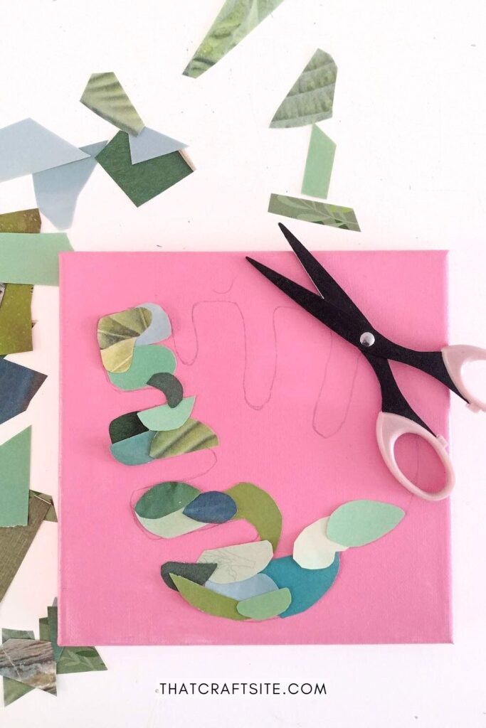 Paper scraps forming the shape of a monstera leaf on pink canvas with scissors next to it.