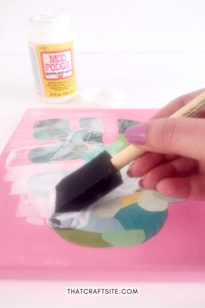 Hand holding foam brush, adding layer of mod podge on Monstera leaf shaped paper collage.