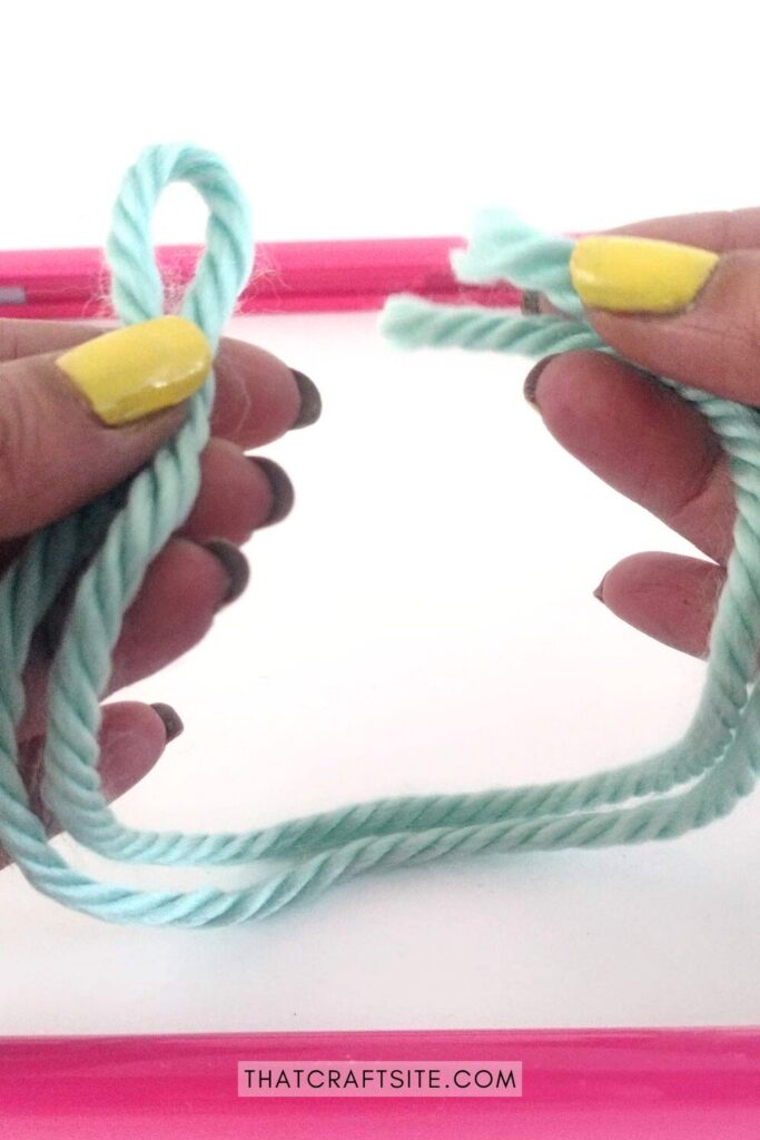 Hands holding up cut piece of blue yarn in front of empty pink frame.